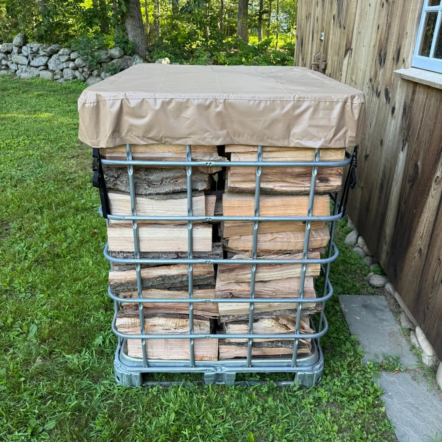 Stack of firewood inside a IBC tote with a cordwood cover tarp on top in an outdoor setting.