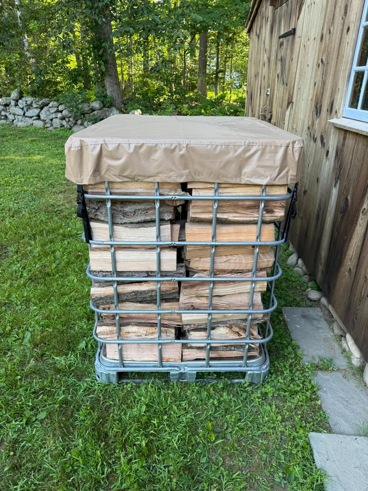 Stack of firewood inside a IBC tote with a cordwood cover tarp on top in an outdoor setting.