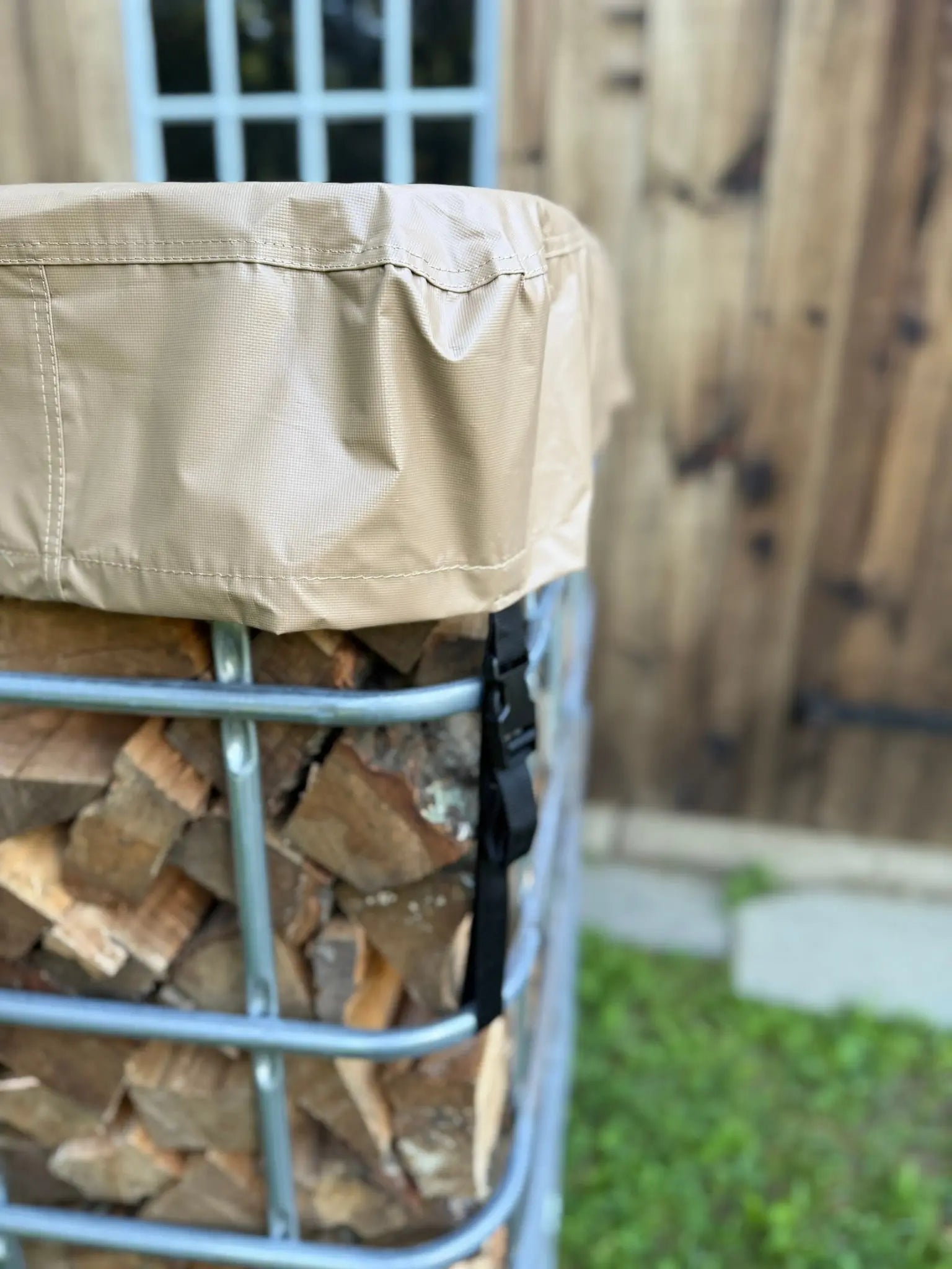 Tan firewood cover on a metal IBC tote firewood rack with stacked firewood inside, against a wooden barn background.