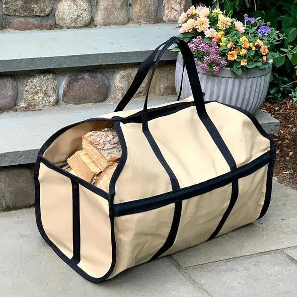 A beige canvas firewood carrier with black trim and handles, filled with logs, placed on a stone step with a potted plant in the background.