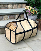 A beige canvas firewood carrier with black trim and handles, filled with logs, placed on a stone step with a potted plant in the background.