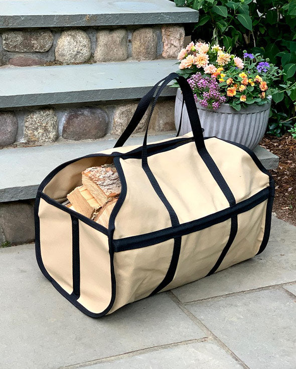 A beige canvas firewood carrier with black trim and handles, filled with logs, placed on a stone step with a potted plant in the background. 
